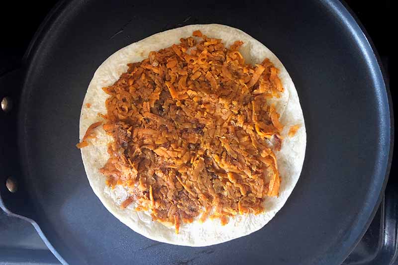Horizontal overhead image of a flour tortilla topped with a shredded and spiced sweet potato mixture in a large nonstick skillet.