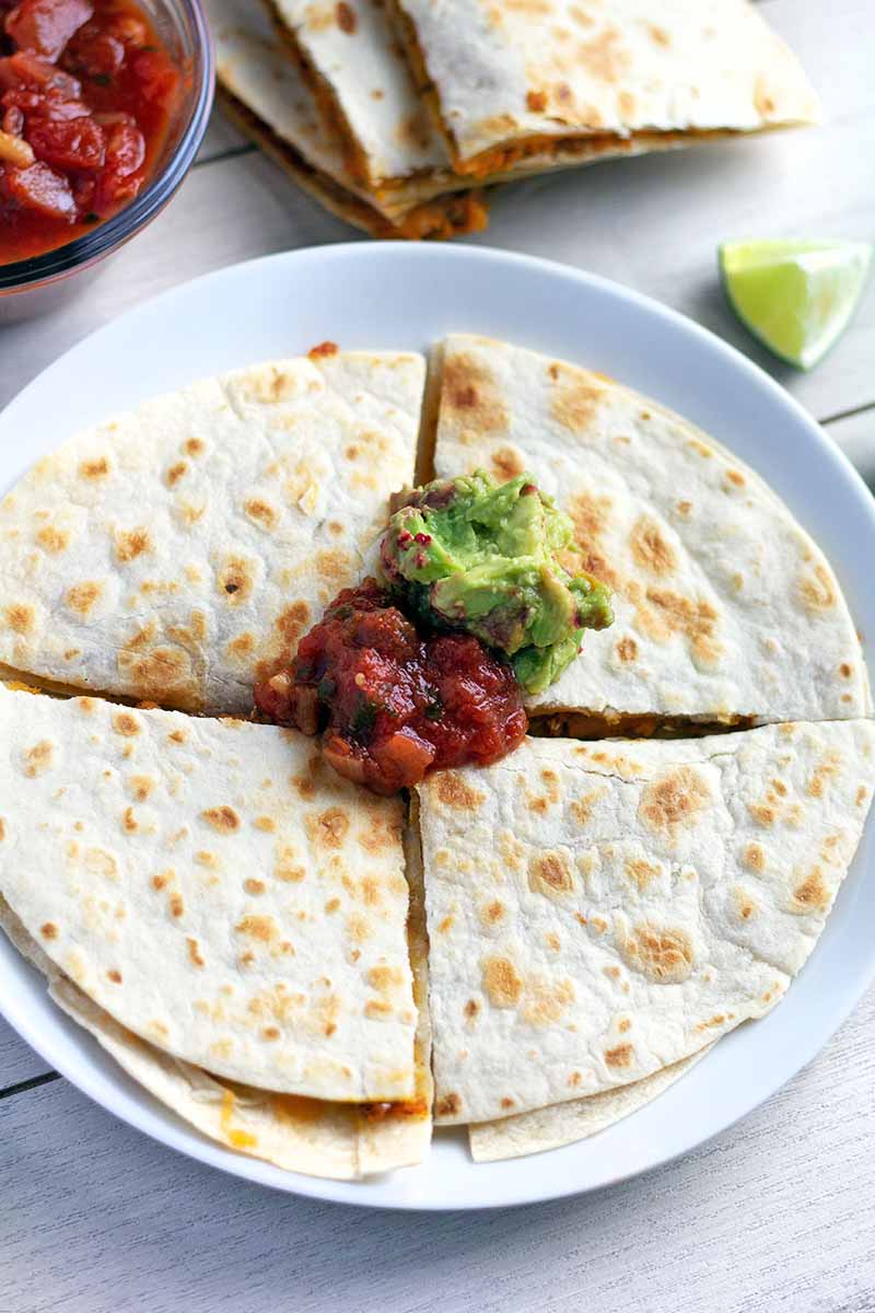 Vertical overhead closely cropped image of a sweet potato quesadilla cut into quarters on a white ceramic plate, topped with salsa and guacamole, on a white wood surface with a glass dish of the red sauce, more pieces of the dish arranged in a stack, and wedges of lime.