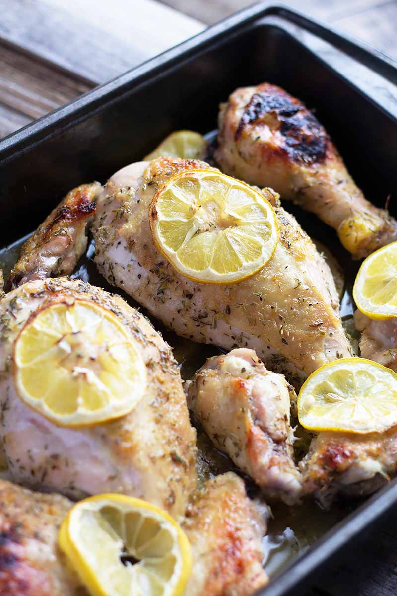 Vertical close-up image of cooked poultry drumsticks topped with thin slices of lemon in a dark roasting pan.