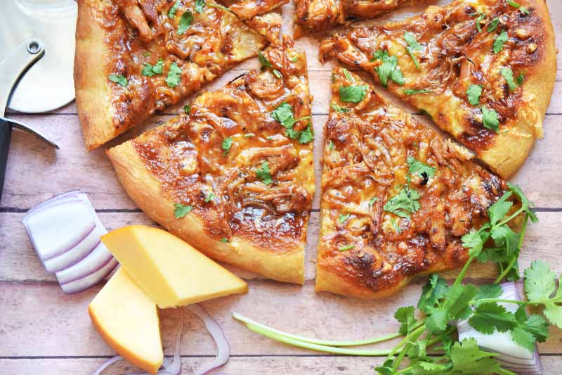 Horizontal overhead closely cropped image of a homemade barbecue chicken pizza that has been cut into triangular slices, on a wood table with a pizza cutting wheel, chunks of gouda cheese, chopped red onion, and sprigs of fresh cilantro.