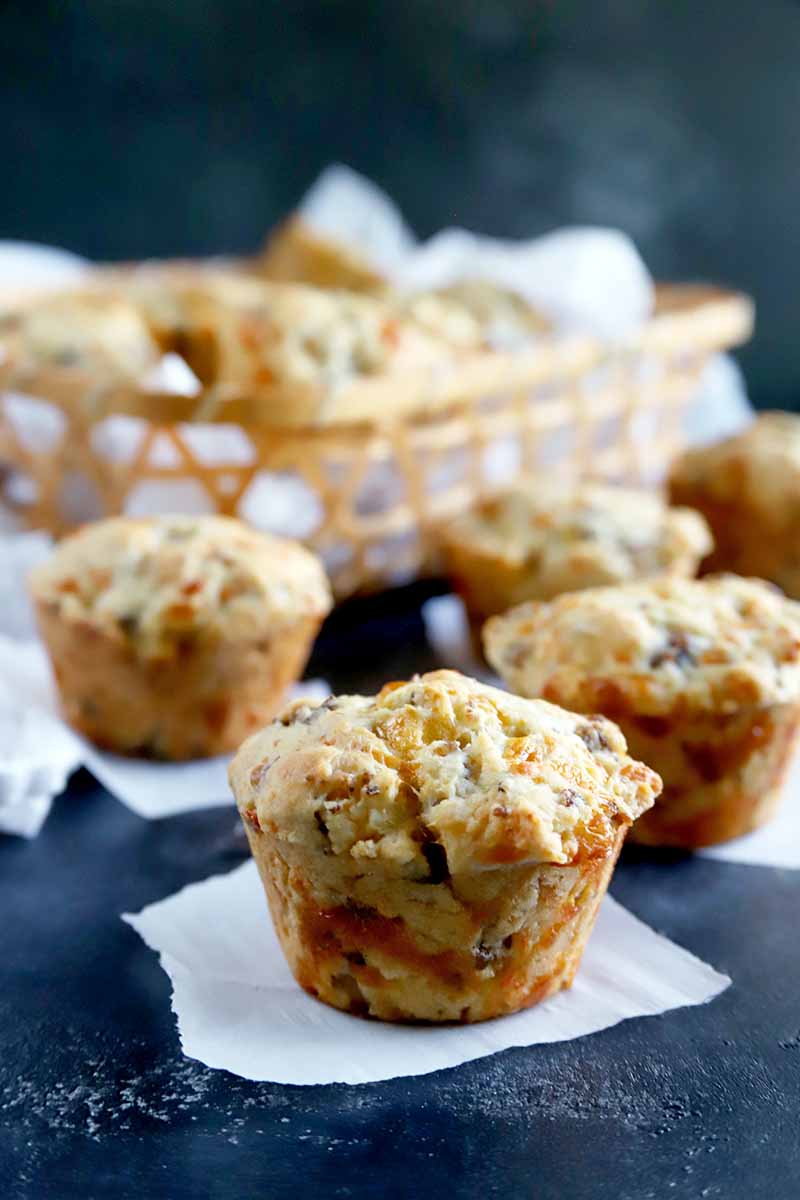 Vertical image of several sausage cheese biscuits that have been baked in muffin tins, arranged on squares of white parchment paper on a dark blue-gray surface, with more in soft focus in a basket in the background.
