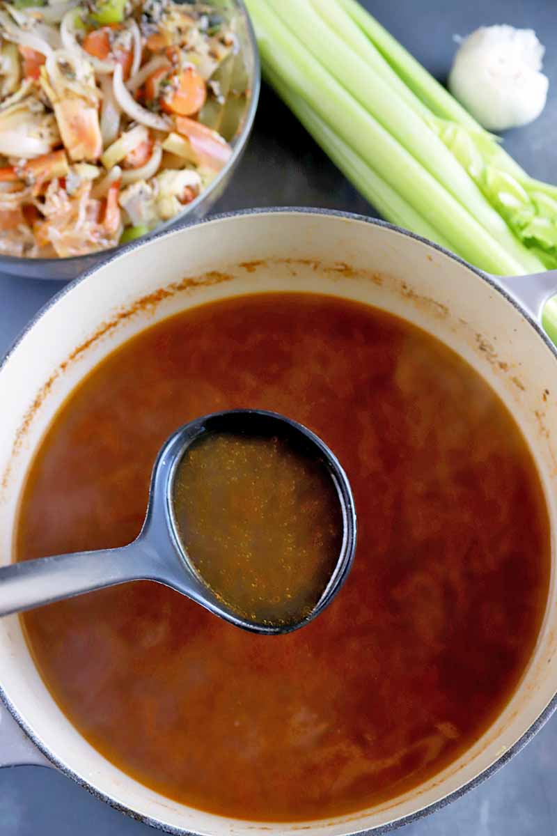 Vertical top-down image of a ladle in a pot with a dark red broth next to celery and other scraps.