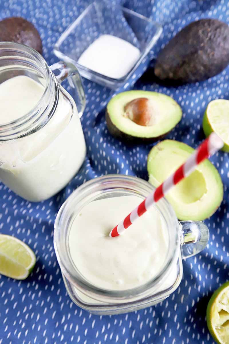 Vertical overhead image of two glass jars filled with a homemade smoothie, with a red and white striped straw, on a blue cloth with white speckles, with lime wedges and cut and whole fresh avocados, and a small square dish of hydrolyzed collagen powder.