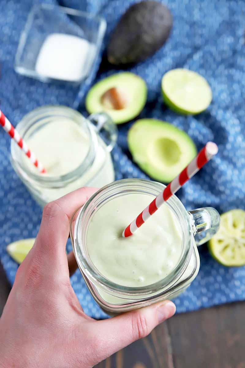 Vertical overhead image of a hand holding a glass drinking vessel with a handle, filled with a pale green smoothie with a red and white striped straw in it, with another identical jar in soft focus in the background, on a blue cloth with a square glass dish of coconut milk, whole and cut avocados, and juiced and sliced limes, on a dark brown wood table.