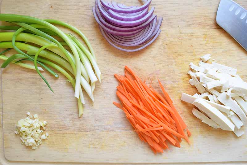 Horizontal image of thinly sliced and minced assorted vegetables and tofu on a wooden cutting board next to a knife.