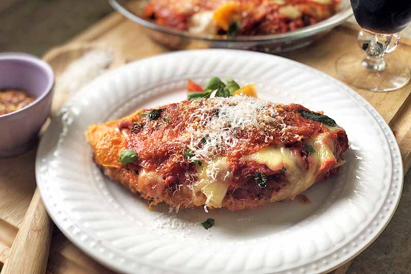 Horizontal oblique overhead image of chicken parmesan with pepperoni on a white plate, garnished with grated parmesan cheese, on a wood surface with a glass of wine, a pie plate containing more of the dish, a small white cup of red pepper flakes, and a wooden cooking spoon.