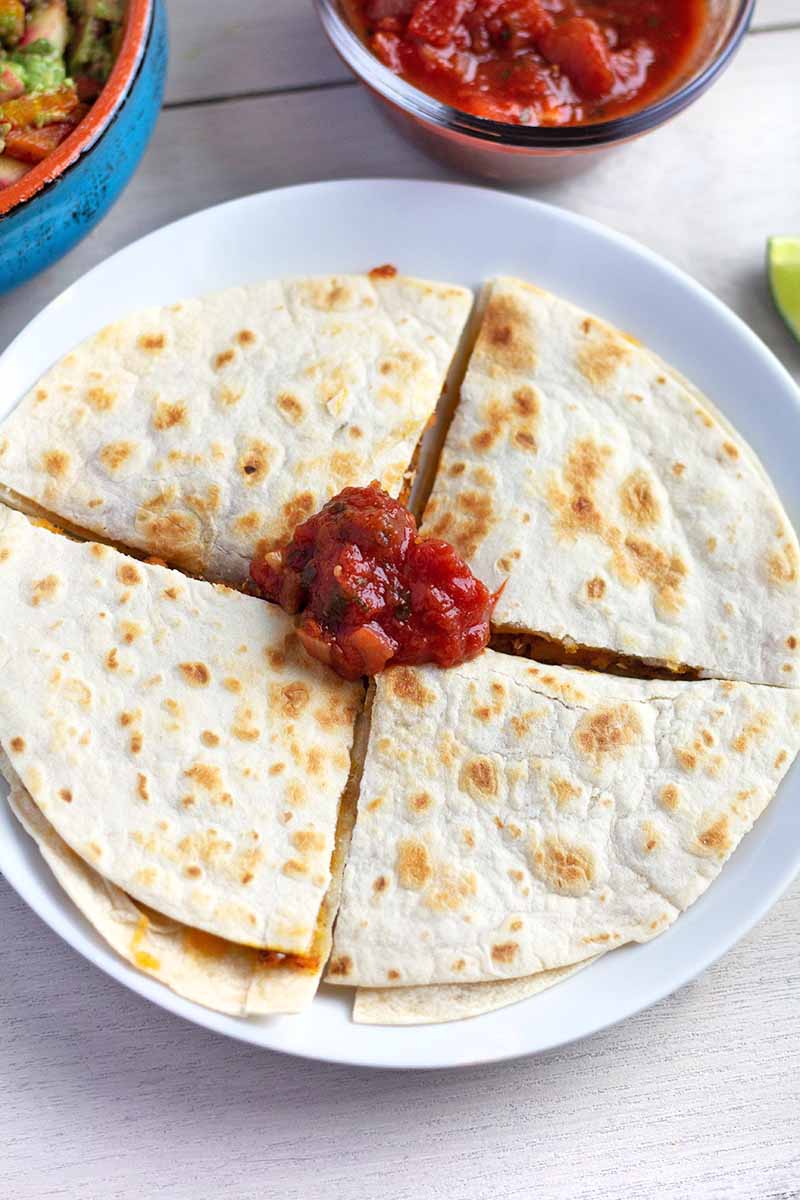 Vertical overhead closely cropped image of a quesadilla that has been cut into four pieces and arranged on a white ceramic plate, topped with a dollop of red tomato salsa, with more of the sauce in a glass bowl at the top of the frame to the right of a blue and orange ceramic bowl of guacamole.