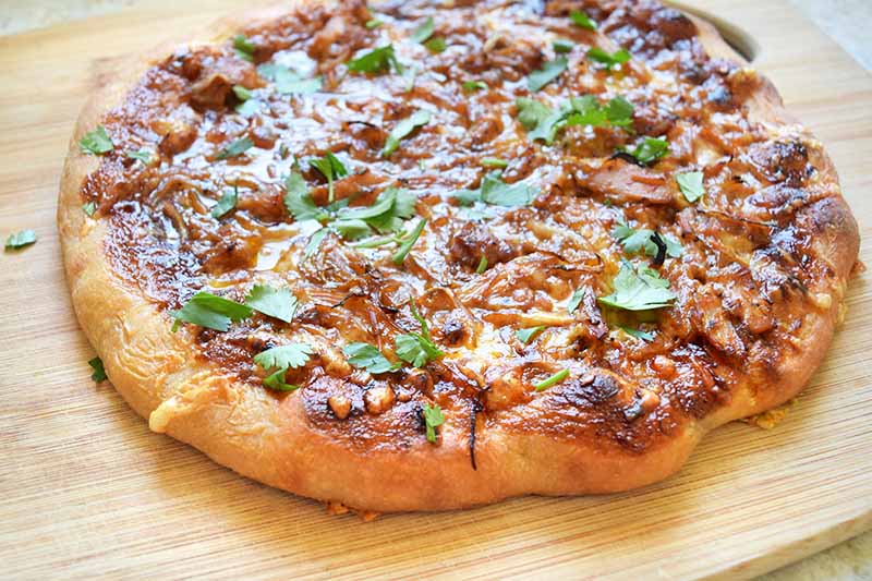 Horizontal image of a freshly baked homemade individually sized pizza topped with grilled poultry, barbecue sauce, red onion, gouda, and fresh cilantro, on a wood cutting board.