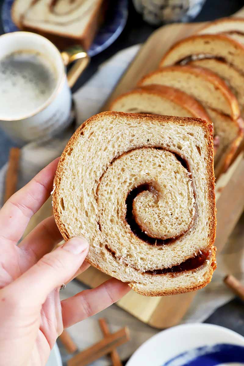 Vertical image of a hand holding a pice of cinnamon swirl bread over a wooden board and mugs of coffee.