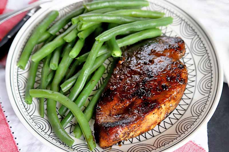 Horizontal overhead image of a portion of balsamic chicken breast and steamed green beans on a white plate with a black patter, on a gray surface topped with a red and white cloth.