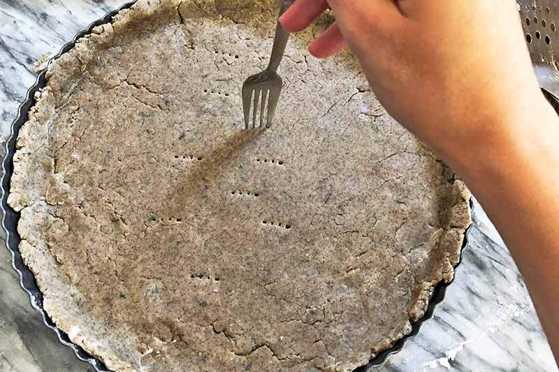 A hand holds a fork and uses it to poke holes in a buckwheat tart crust that has been arranged in a metal pan, on a gray and white marble surface.