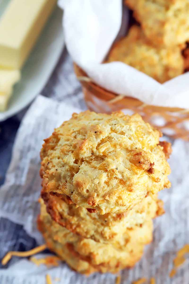 Overhead vertical image of a stack of three cheddar garlic biscuits next to a basket containing more, with a white ceramic dish of butter on pieces of tissue paper, with scattered shredded cheese bits.