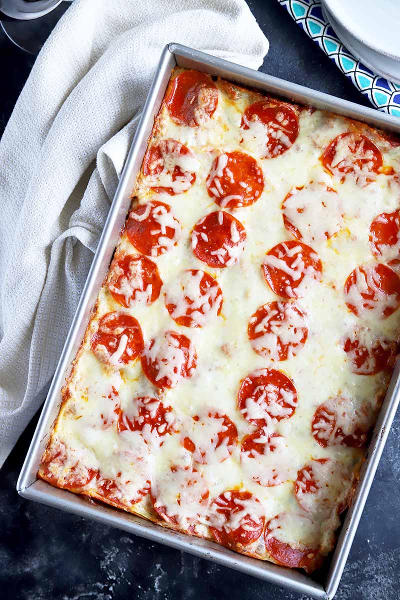 Overhead horizontal image of a rectangular metal baking pan of lasagna topped with pepperoni and melted cheese, with a folded white cloth on a gray surface.