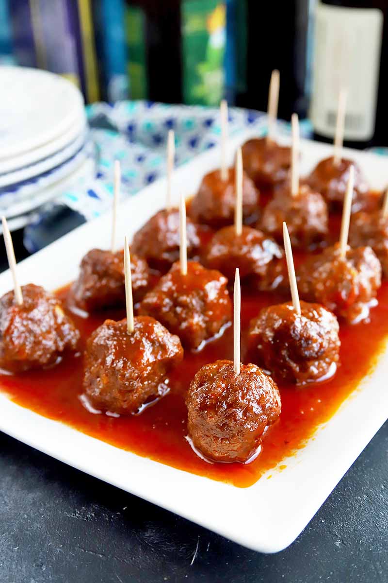 Vertical image of a white platter with neat rows of meatballs with toothpicks covered in a red sauce.