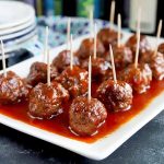 Horizontal image of a white platter with rows of meatballs covered in a red sauce with toothpicks inserted in them.