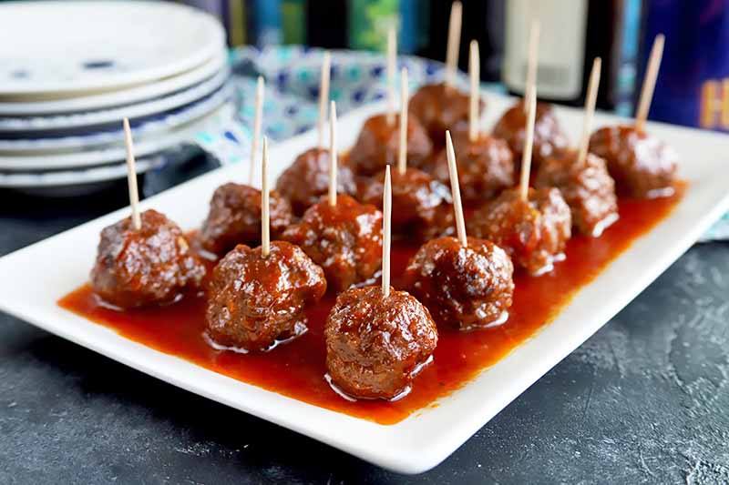 Horizontal image of a white platter with rows of meatballs covered in a red sauce with toothpicks inserted in them.