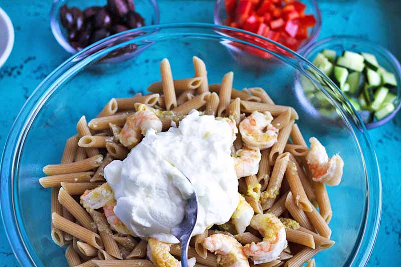 Closely cropped horizontal overhead image of a large glass mixing bowl of cooked whole wheat penne with shrimp, topped with a dollop of Greek yogurt, surrounded with small glass bowls of chopped olives, red bell pepper, and cucumber, on a blue surface.