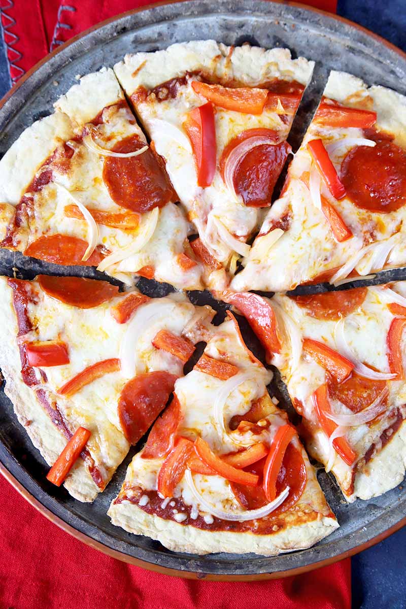 Vertical overhead closely cropped image of a pepperoni, red bell pepper, and onion pizza on an einkorn crust on a metal pan, on a gray surface topped with a red cloth.