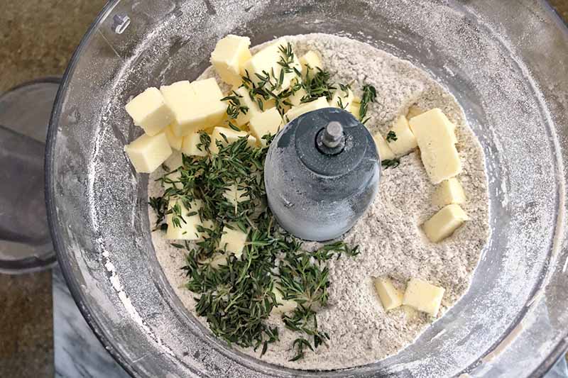 Closely cropped horizontal overhead image of buckwheat flour, fresh thyme, and cubes of chilled butter in a food processor, on a brown surface.