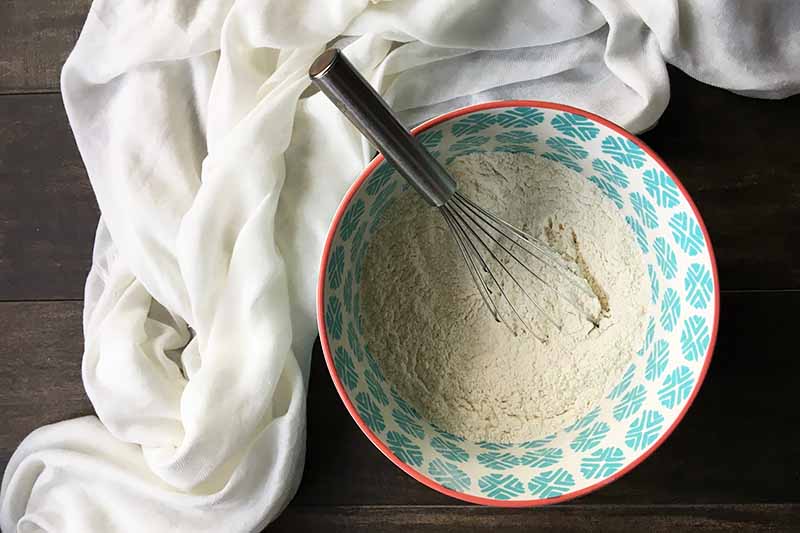 Horizontal image of a blue bowl with dry ingredients and a whisk next to a white towel.