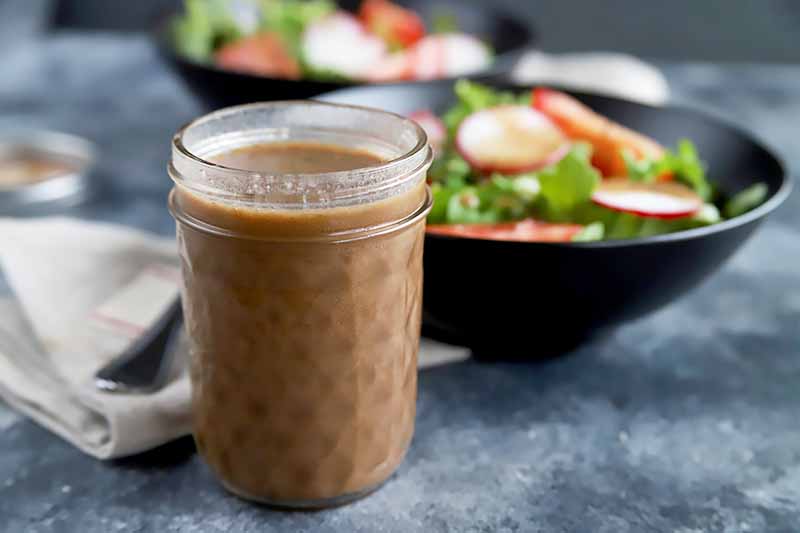 Horizontal image of a glass jar with a brown creamy liquid in front of a napkin, fork, and bowls of salad.