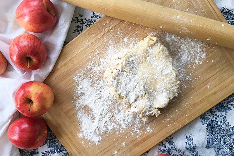 Horizontal image of a thick dough on a wooden cutting board covered in flour next to a rolling pin.
