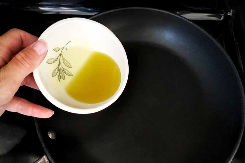 Horizontal image of a hand about to pour a small white ceramic bowl of olive oil into a large nonstick frying pan.