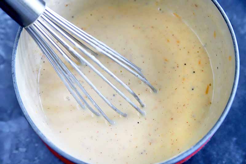 Closely cropped close-up overhead horizontal image of a silver metal whisk stirring a cheese sauce in an enameled pan, on a mottled blue-gray surface.