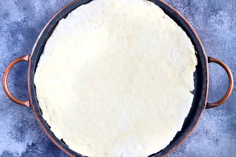 Horizontal closely cropped overhead image of a rolled out round portion of pizza dough on a metal baking pan with two handles, on a sponge painted glue and white surface.