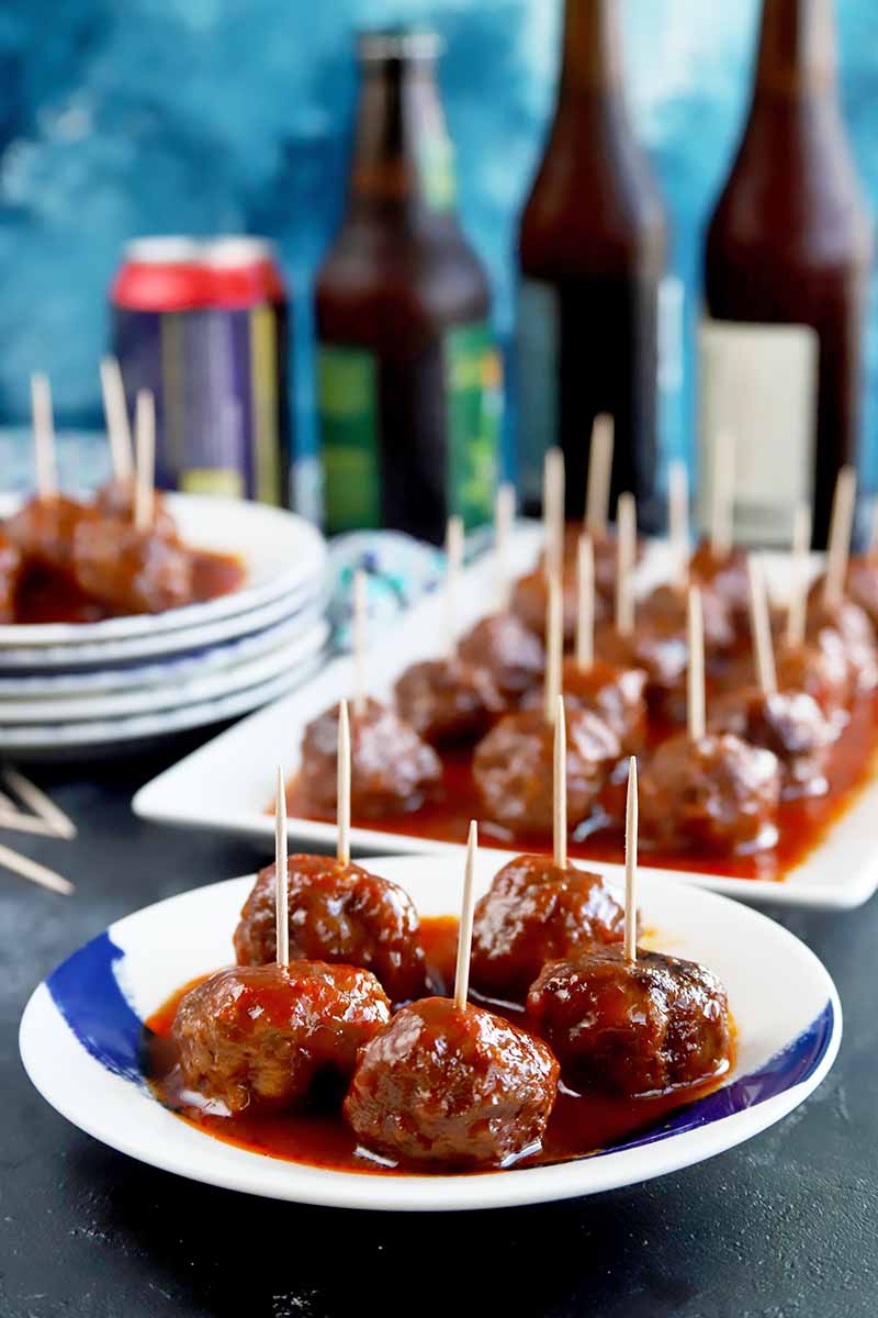 Vertical image of plates and platters with meatballs in sauce with toothpicks inserted in them, in front of bottles and a can.