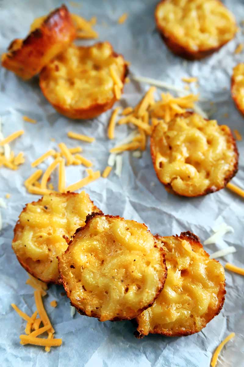 Vertical close-up image of elbow macaroni with cheese sauce that has been baked in a mini muffin pan, on a crumpled piece of white parchment paper with scattered shreds of cheddar.