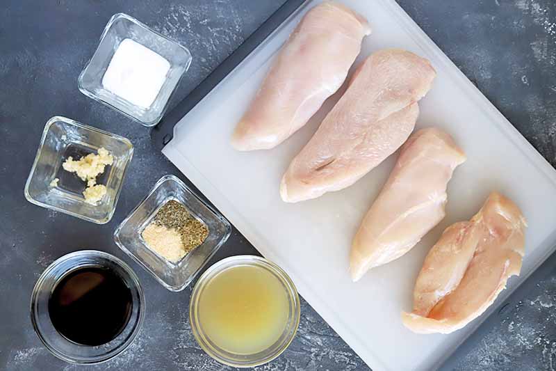 Horizontal overhead closely cropped image of four chicken breasts on a white plastic cutting board with small square and round glass bowls of sugar, spices, minced garlic, chicken broth, and balsamic vinegar, on a gray surface.