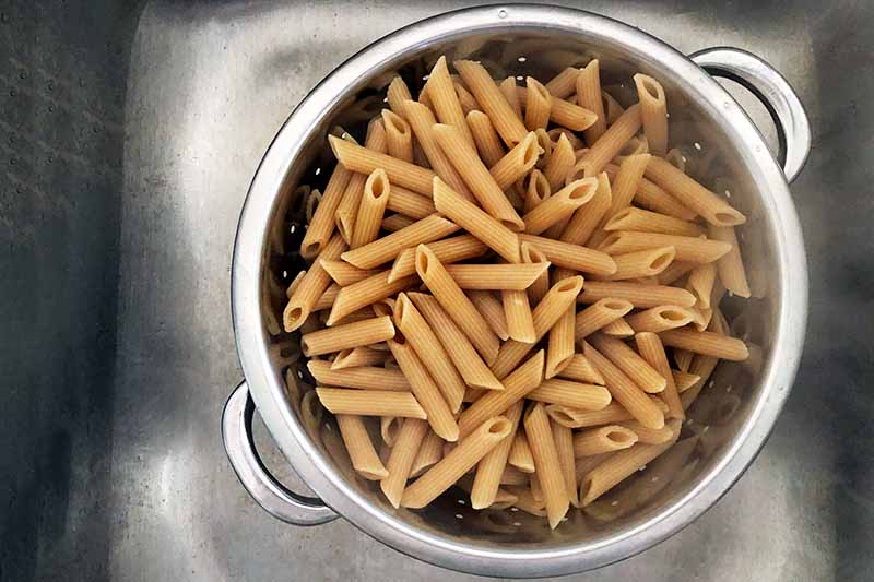 Overhead horizontal image of cooked whole wheat penne draining in a colander in a metal kitchen sink.