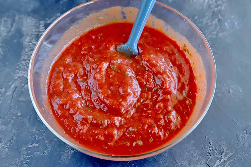 Overhead image of a glass mixing bowl of red marinara sauce being stirred with a blue silicone spatula, on a blue-gray surface.
