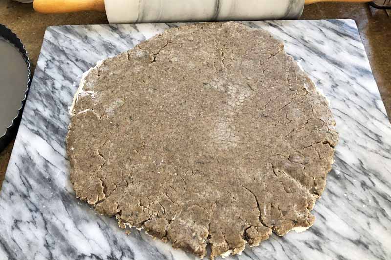 Horizontal image of a rolled out circle of buckwheat pie dough on a marble slab, with a marble and wood rolling pin at the top of the frame.