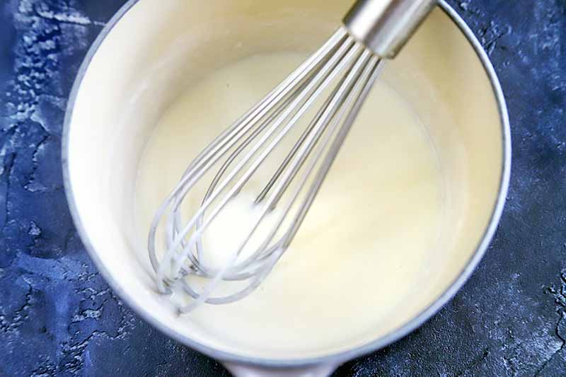 Horizontal closely cropped overhead image of a wire whisk stirring a cream sauce in an enameled saucepan, on a blue-gray sponge-painted surface.