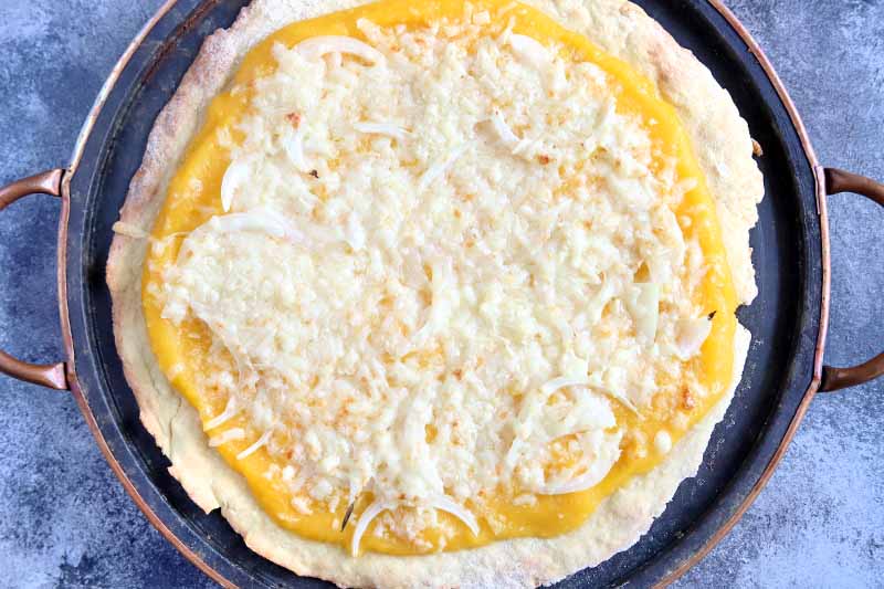 Horizontal overhead image of a rolled out round of pizza dough topped with squash puree, onions, and cheese, on a round metal baking pan with two handles, on a sponge-painted blue-gray and white surface.