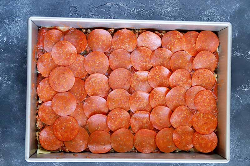 Horizontal overhead image of slices of pepperoni arranged in a slightly overlapping single layer in a large rectangular metal baking pan, on a white-speckled blue-gray painted surface.