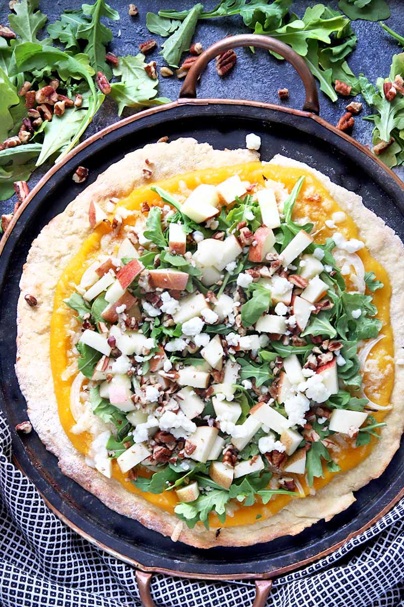 Vertical overhead image of a homemade pizza topped with arugula, cheese, nuts, and chopped apples, on a squash puree base, on a metal baking pan with two handles, on a gray surface with a black and white checkered cloth and scattered topping ingredients.