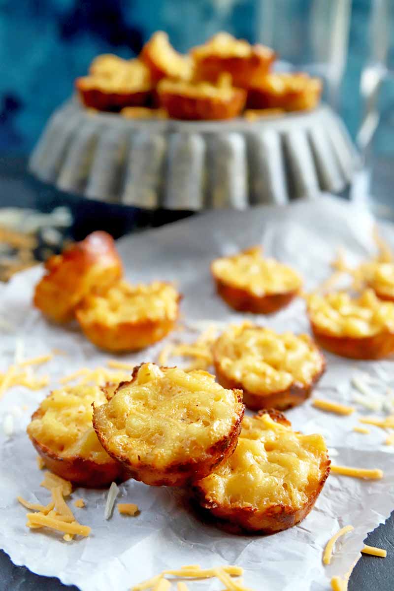 Vertical image of mini baked macaroni bites on a white piece of parchment with scattered pieces of shredded cheddar, and on a gray display pedestal in the background, against a mottled blue backdrop.