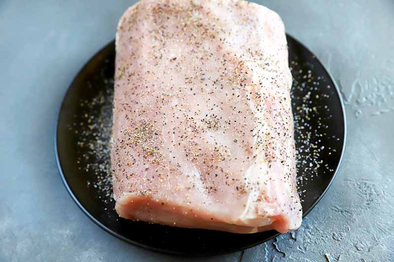 Horizontal oblique overhead image of raw pork seasoned with salt and pepper on a black plate, on a gray surface.