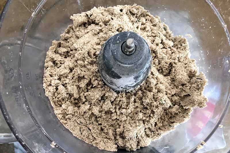Closely cropped overhead horizontal image of a lumpy buckwheat flour mixture in a food processor with a clear plastic canister.