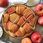 Horizontal image of a whole apple pastry in a pan next to a bowl of flour, whole fruit, and a serving knife.