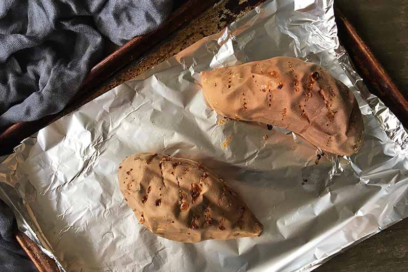 Horizontal image of cooked whole sweet potatoes pricked with fork tines on a baking sheet lined with foil.