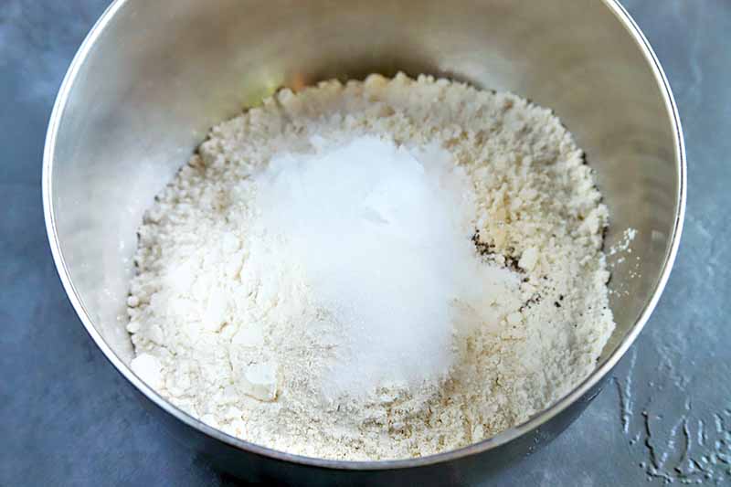 Horizontal overhead closely cropped image of flour and other dry ingredients in a stainless steel mixing bowl, on a gray background.
