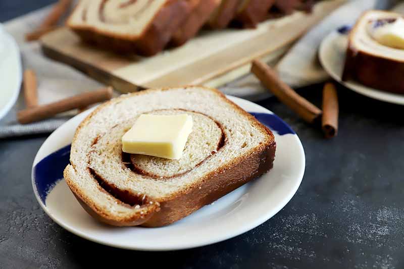 Horizontal image of a slice of swirled bread on a plate next to cinnamon sticks.