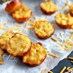 Overhead horizontal image of miniature baked macaroni appetizers on a crumpled piece of white parchment paper, with shredded cheddar cheese scattered on the surface, on a gray table.
