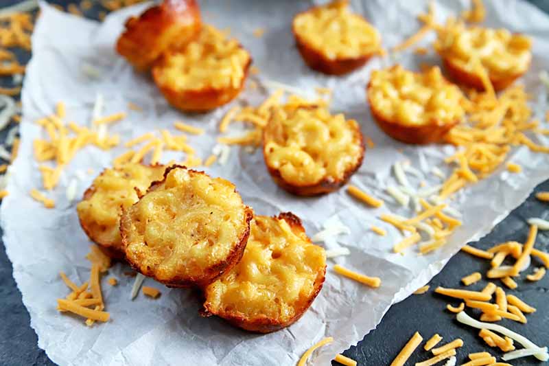 Overhead horizontal image of miniature baked macaroni appetizers on a crumpled piece of white parchment paper, with shredded cheddar cheese scattered on the surface, on a gray table.
