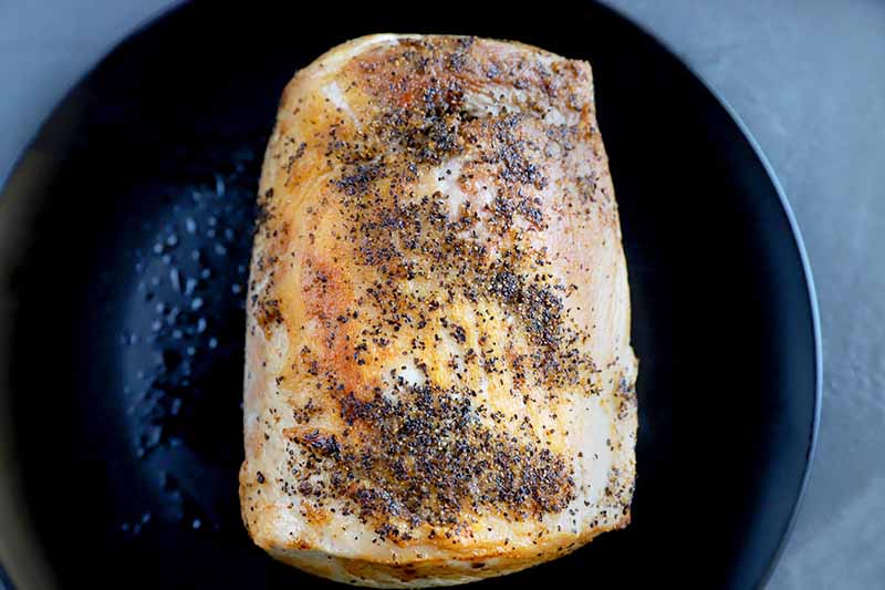 Seasoned pork that has been seared and developed a brown crust on the outside, in a black cast iron pan on a gray surface.