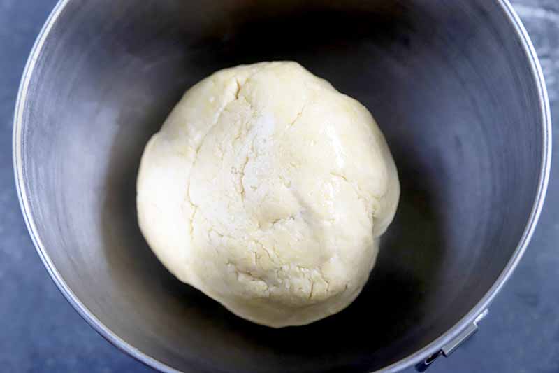 Closely cropped overhead horizontal image of a lightly oiled ball of dough in a stainless steel mixing bowl, on a blue-gray surface.